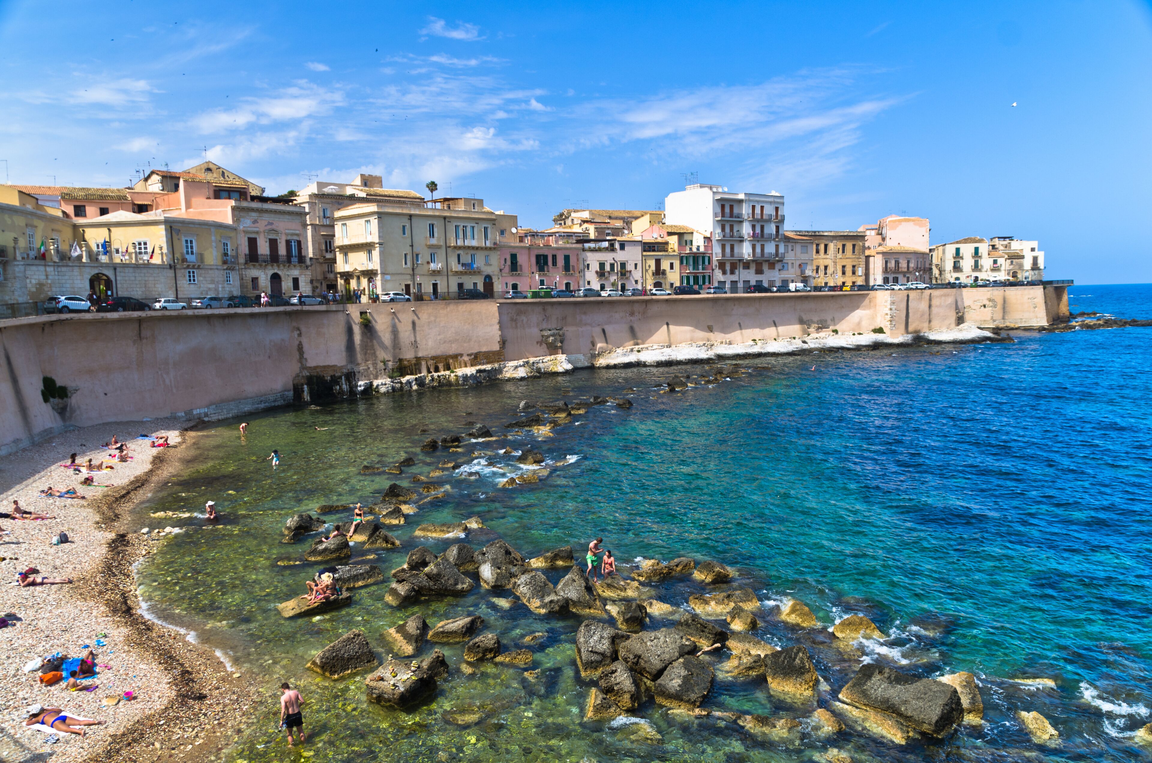 A coastal town with a clear blue sea, rocky shoreline, and historic buildings along the waterfront.