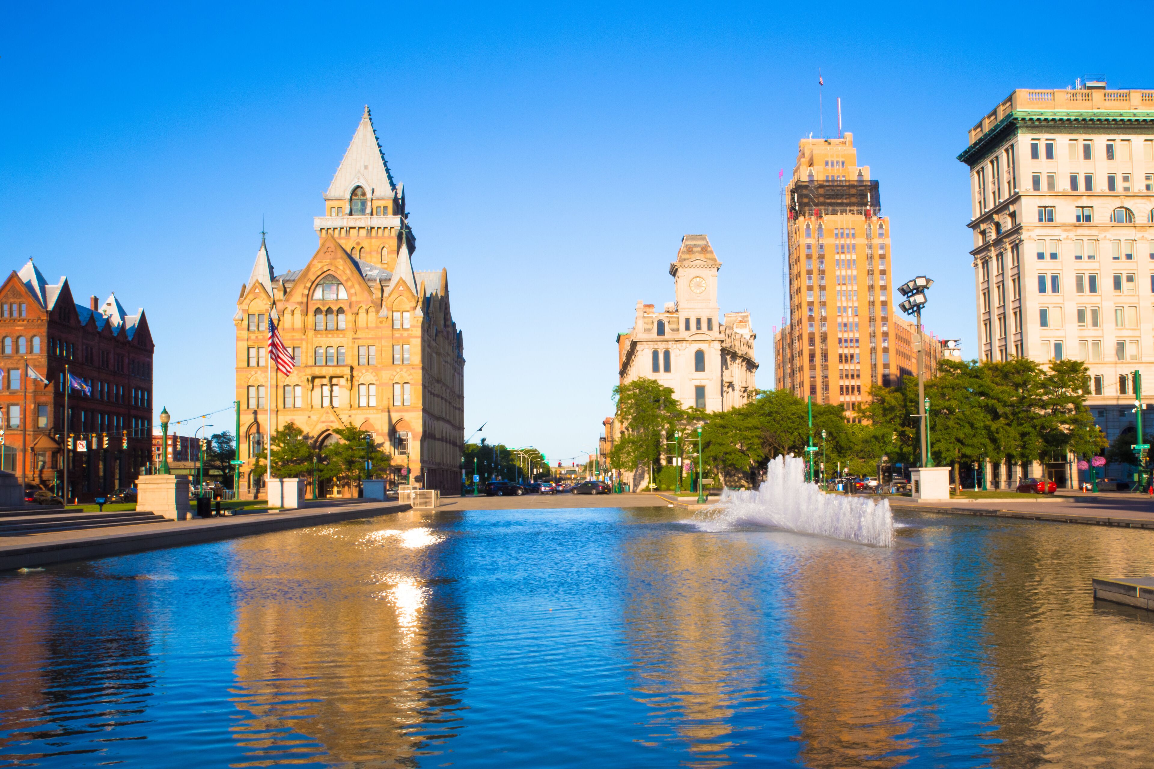 A city square with a fountain, historic buildings, and a clear blue sky.