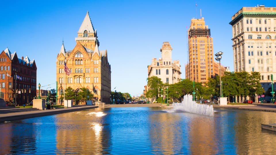 A city square with a fountain, historic buildings, and a clear blue sky.