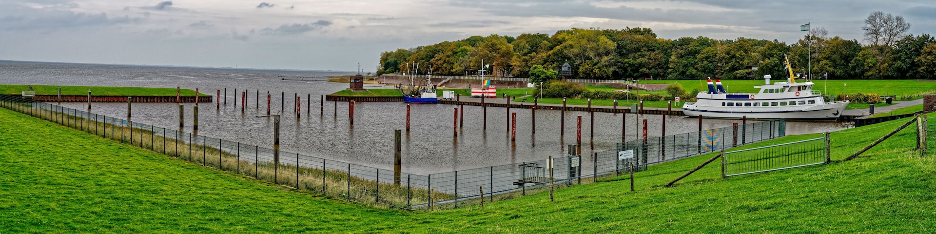A river with a boat docked at a pier, surrounded by green grass and a cloudy sky.
