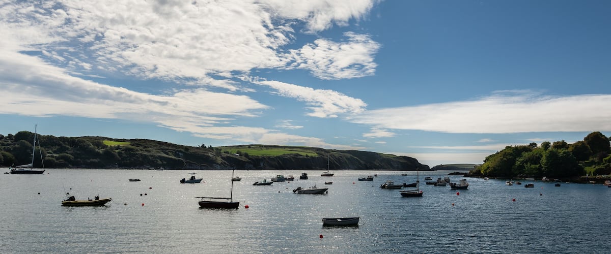 A coastal scene with numerous boats on calm waters, surrounded by green hills and a partly cloudy sky.
