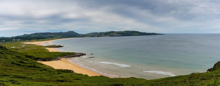 Un paisaje costero con una playa arenosa, colinas verdes y un mar tranquilo.