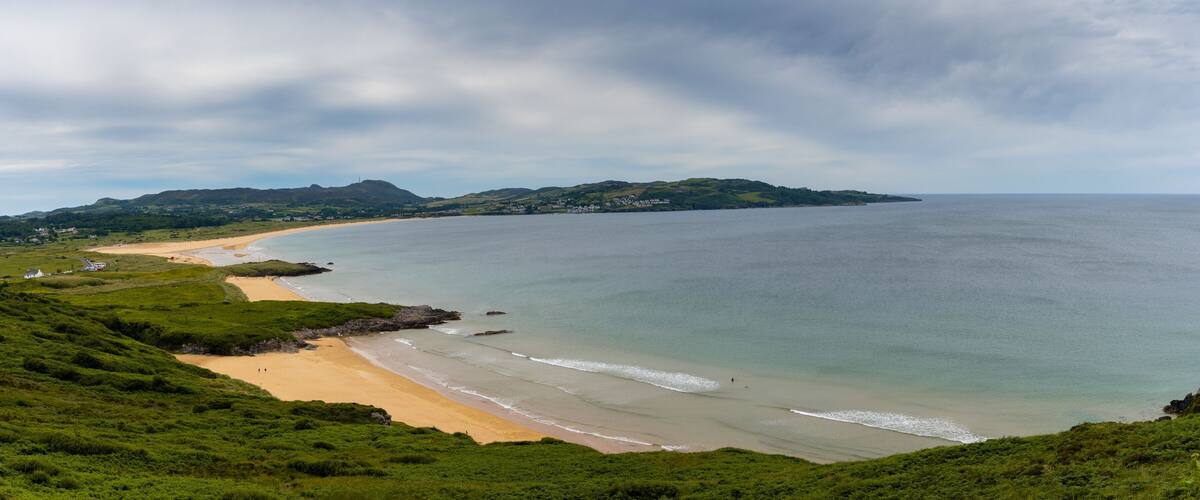 A coastal landscape with a sandy beach, green hills, and a calm sea.