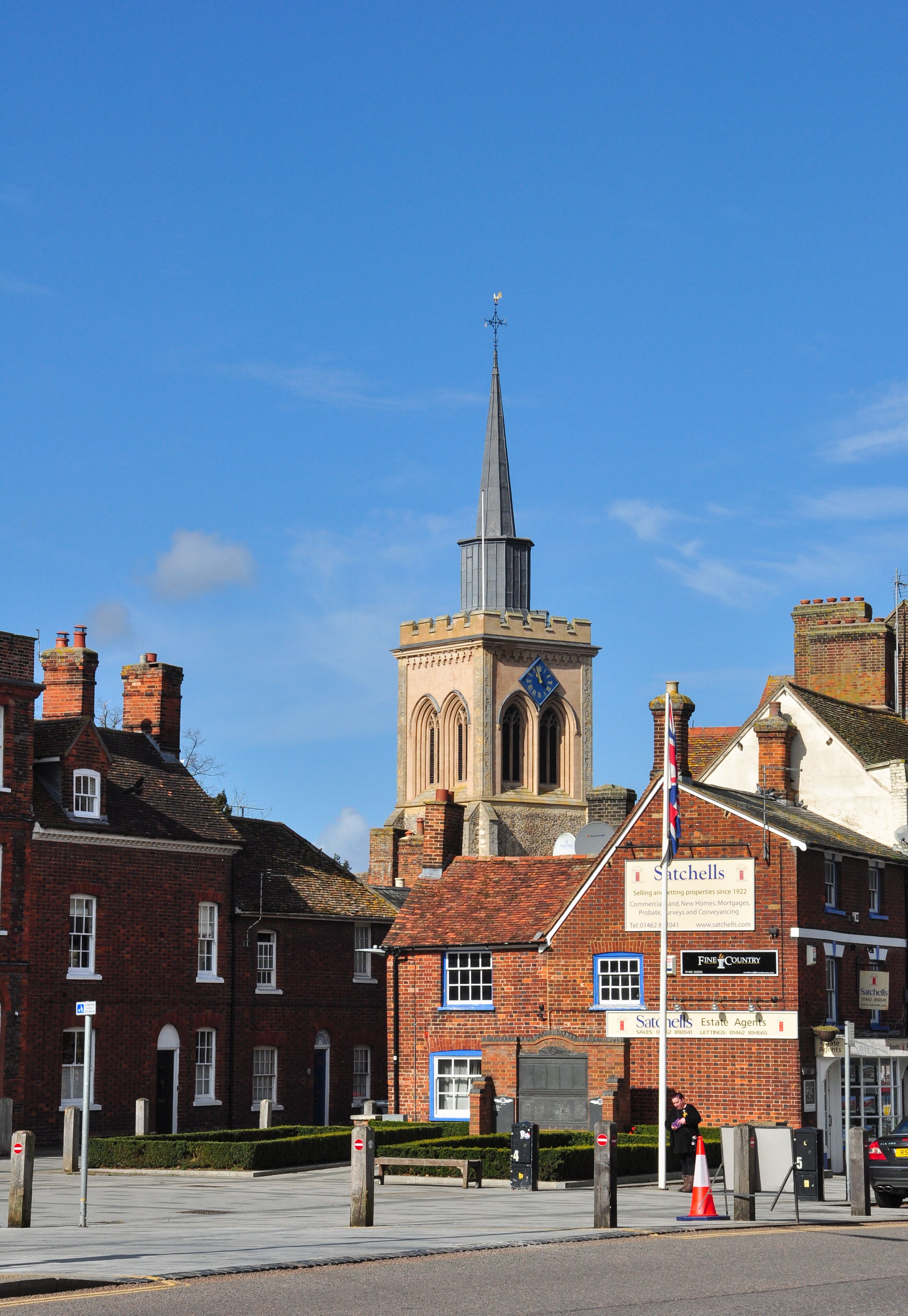 A church with a tall spire, a red brick building with a sign for 'Satchells Estate Agents', and a clear blue sky.
