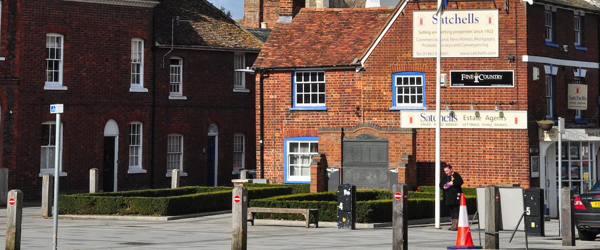 A church with a tall spire, a red brick building with a sign for 'Satchells Estate Agents', and a clear blue sky.