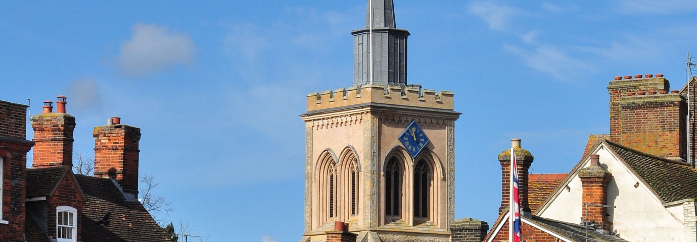 A church with a tall spire, a red brick building with a sign for 'Satchells Estate Agents', and a clear blue sky.
