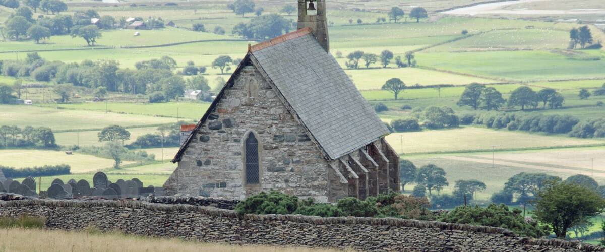 Uma igreja de pedra com um campanário, rodeada por campos verdes e ovelhas a pastar.