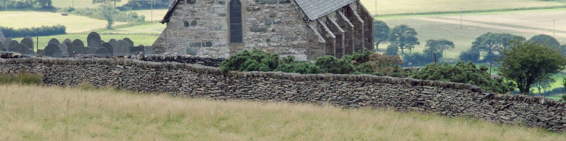 A stone church with a bell tower, surrounded by green fields and sheep grazing.