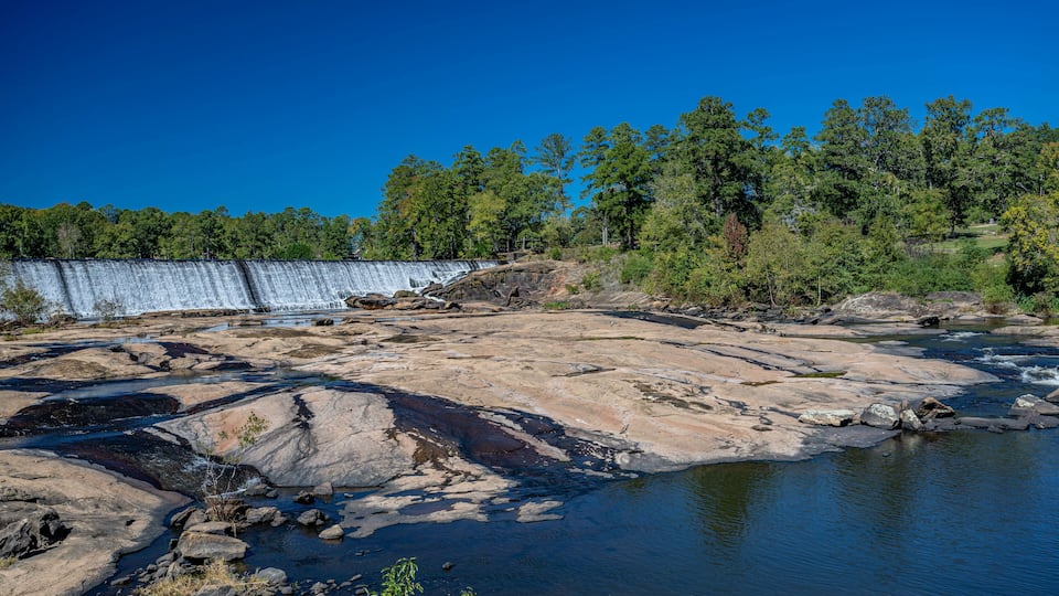 A river flowing through a rocky landscape with a dam in the background and a forested area.