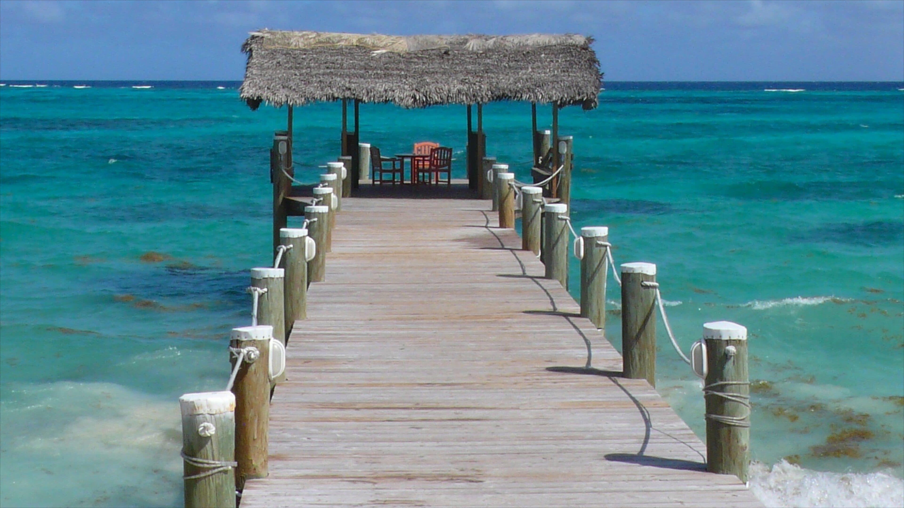 A wooden pier leading to a thatched-roof structure with chairs.