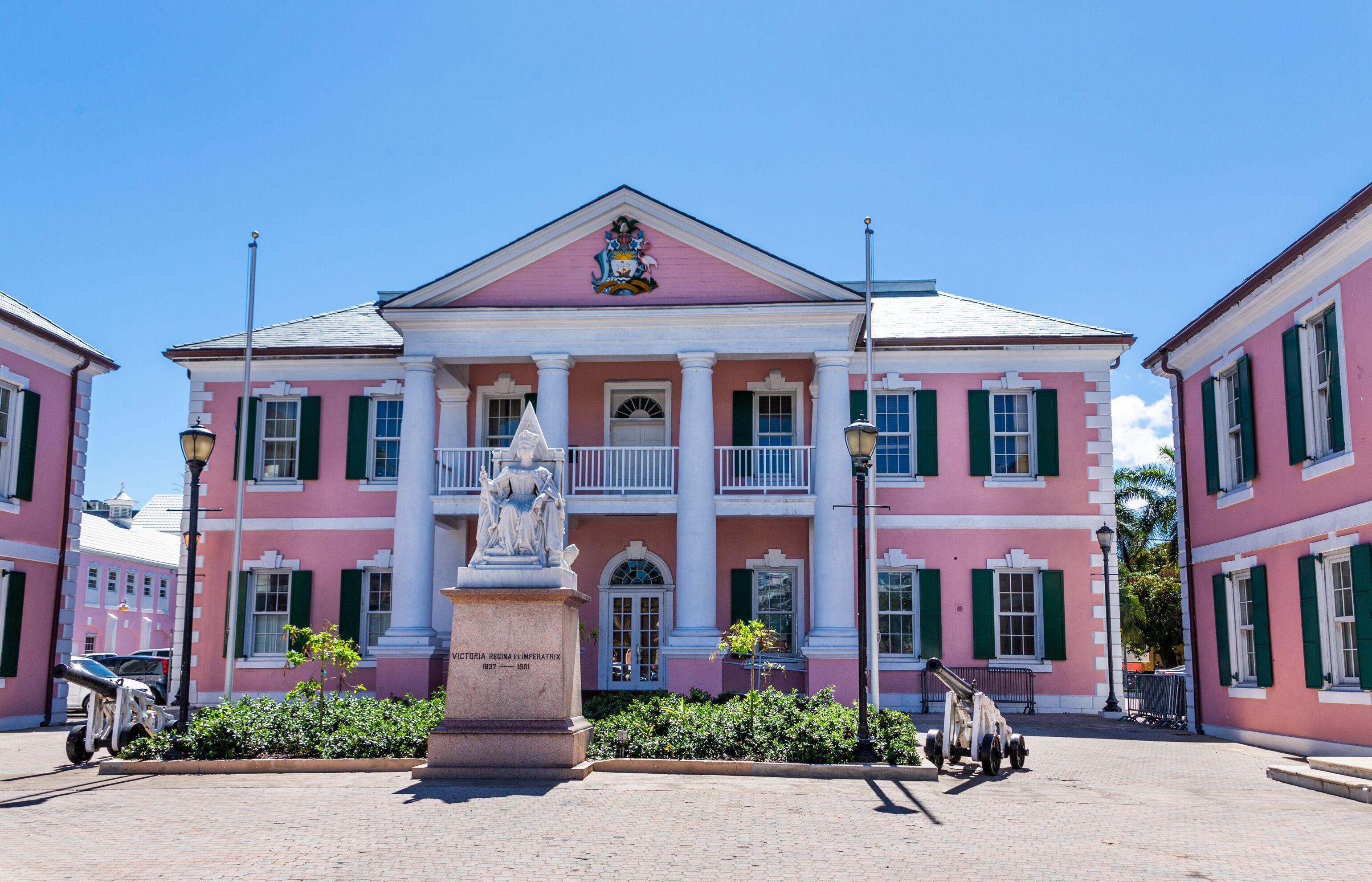 A pink colonial-style building with green shutters and a balcony, a statue in front, and a plaque reading "Victoria Regina Imperatrix 1837-1883".