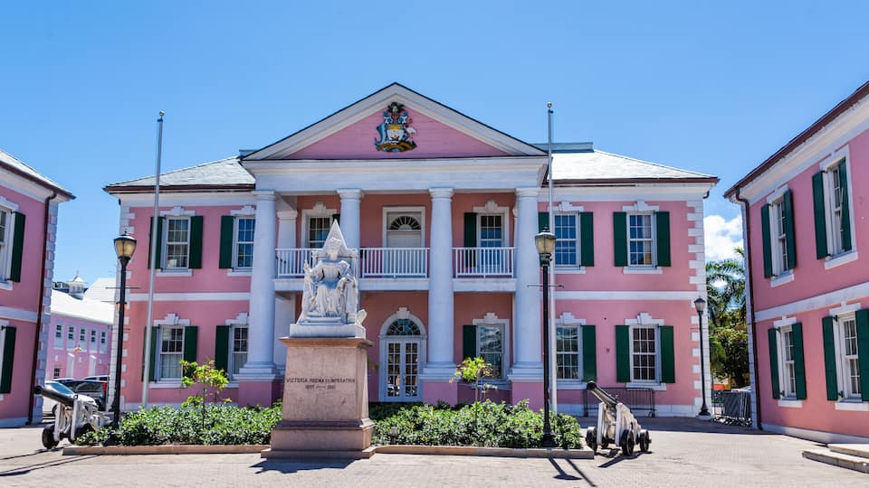 A pink colonial-style building with green shutters and a balcony, a statue in front, and a plaque reading "Victoria Regina Imperatrix 1837-1883".