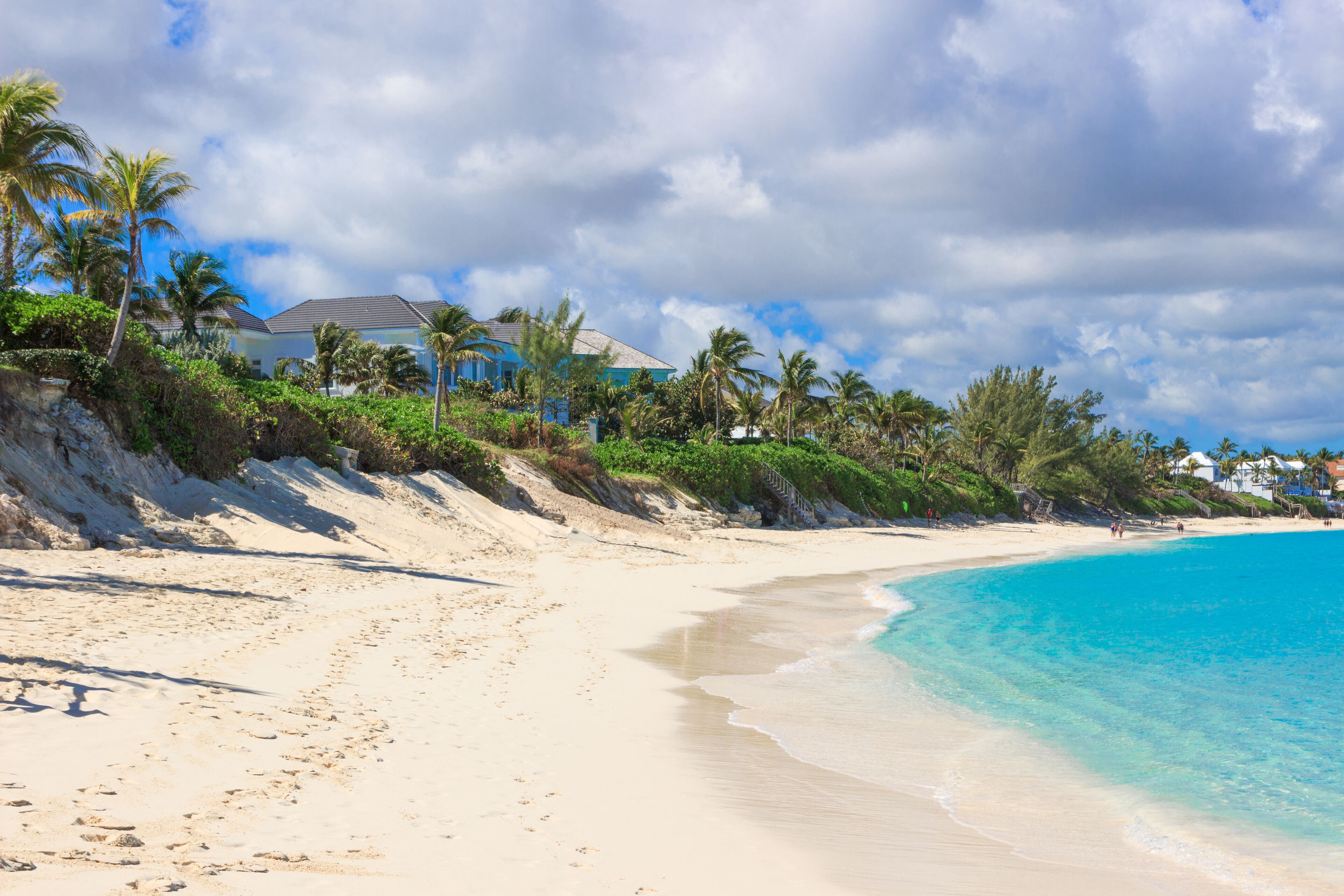 A sandy beach with clear blue water, palm trees, and houses on a hillside.
