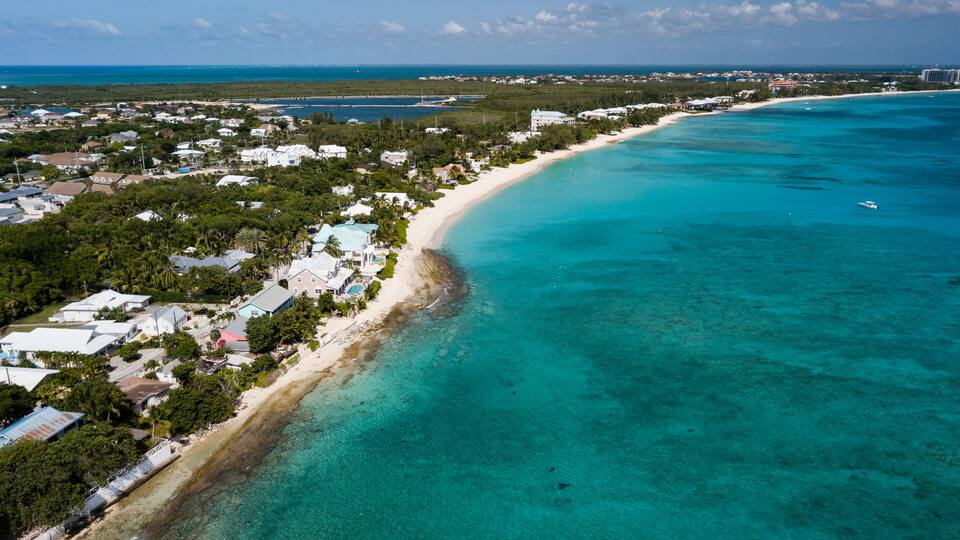 An aerial view of a coastal town with a long sandy beach, clear turquoise waters, and dense greenery.