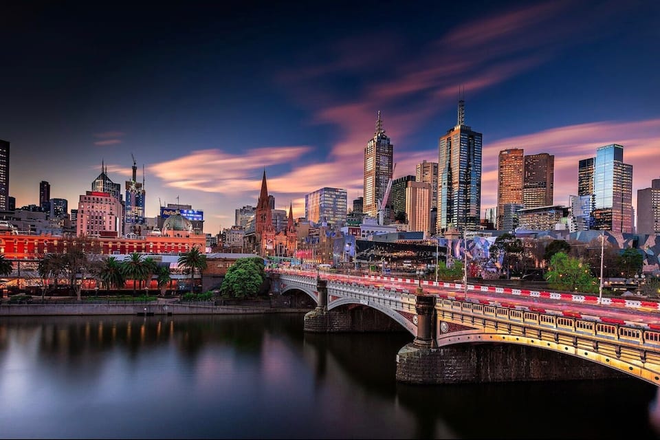 A city skyline at dusk with illuminated buildings, a bridge over a river, and a historic building with a dome.