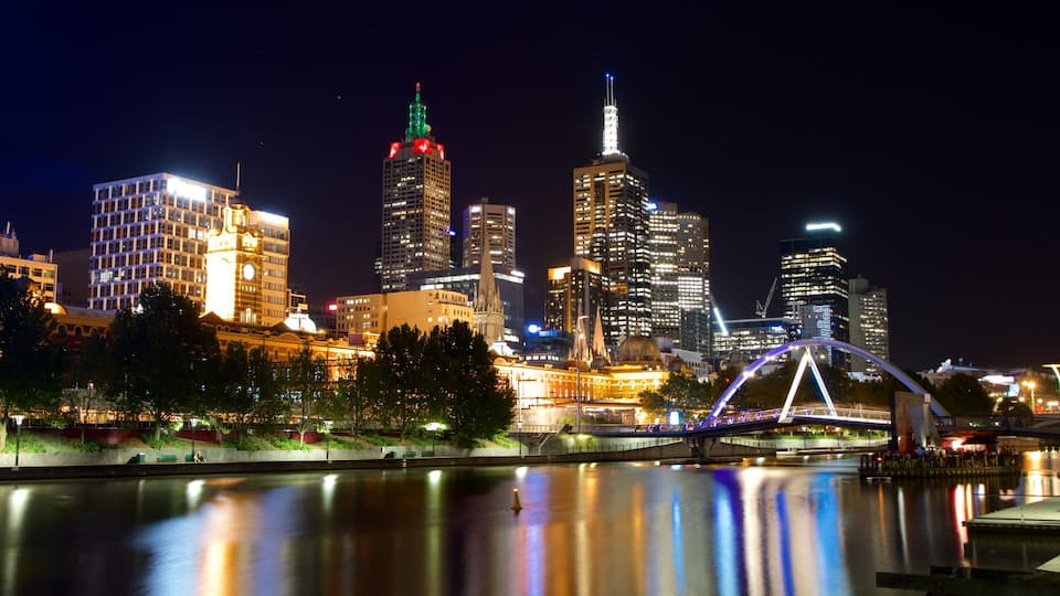 A city skyline at night with illuminated buildings, a river in the foreground, and a bridge with lights.
