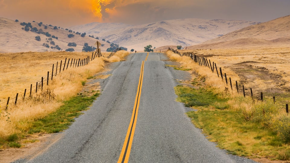 A rural road flanked by wooden fences, leading towards distant hills under a sunset sky.