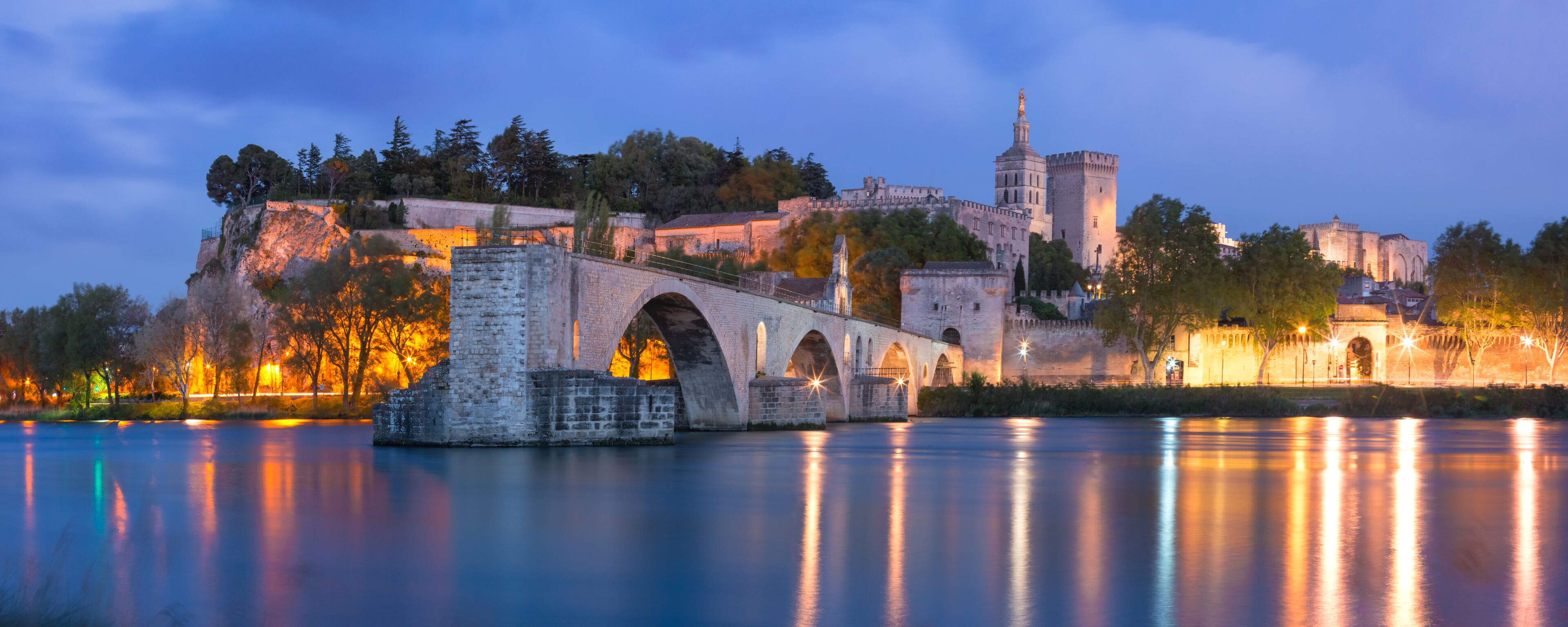 A stone bridge over a calm river, a castle with a tower, and a hill with trees.