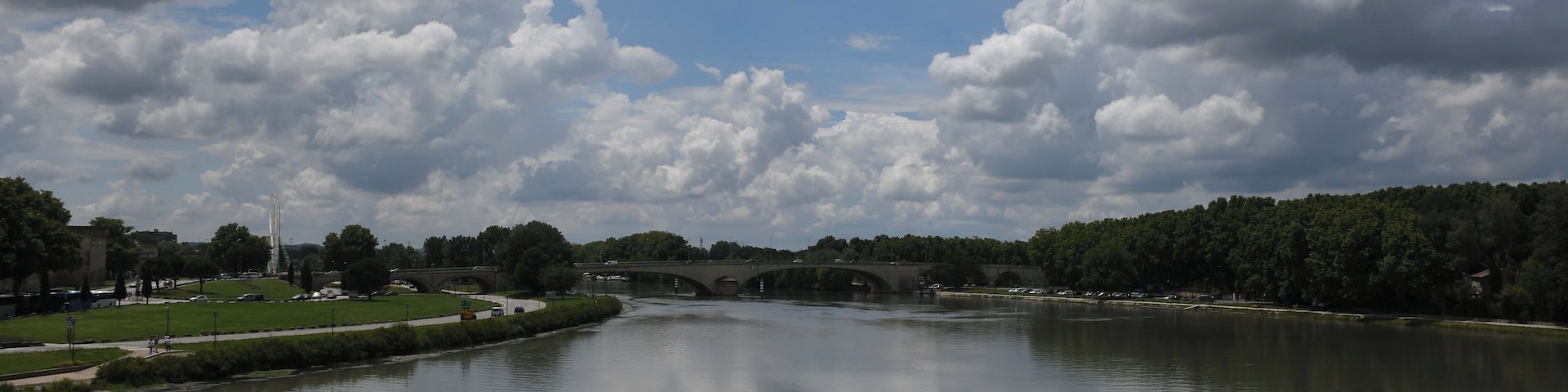 Une rivière traversée par un pont, bordée d’arbres et surplombée d’un ciel dégagé.