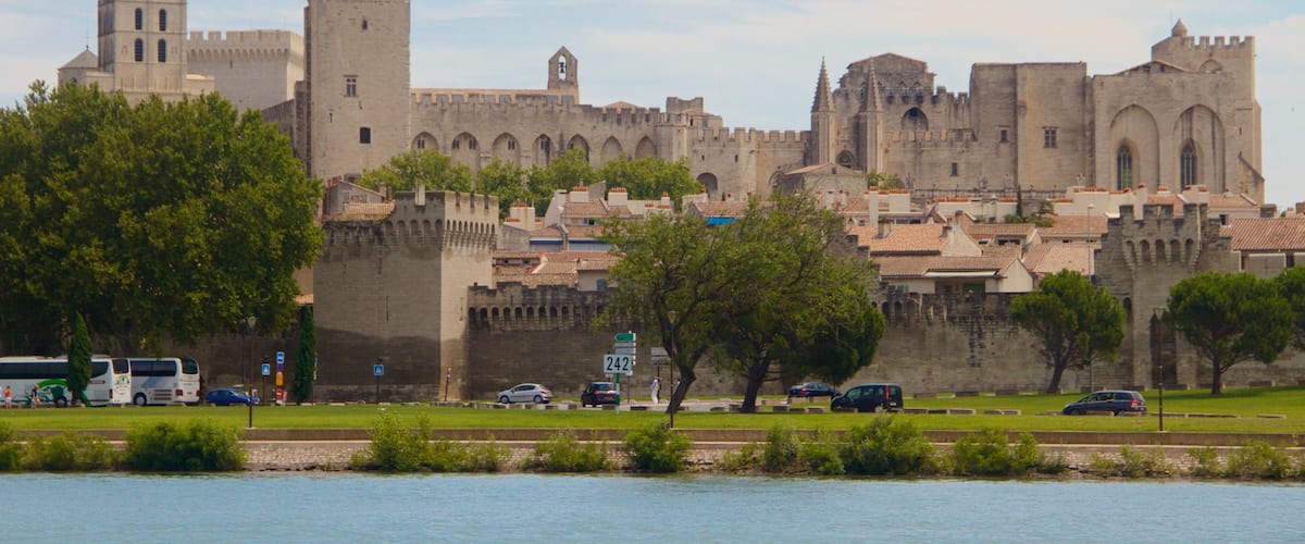 A historic castle with a prominent tower and a statue on top, overlooking a river with a bridge and greenery in the foreground.