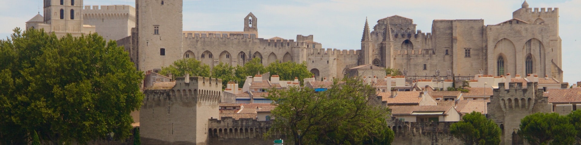 A historic castle with a prominent tower and a statue on top, overlooking a river with a bridge and greenery in the foreground.