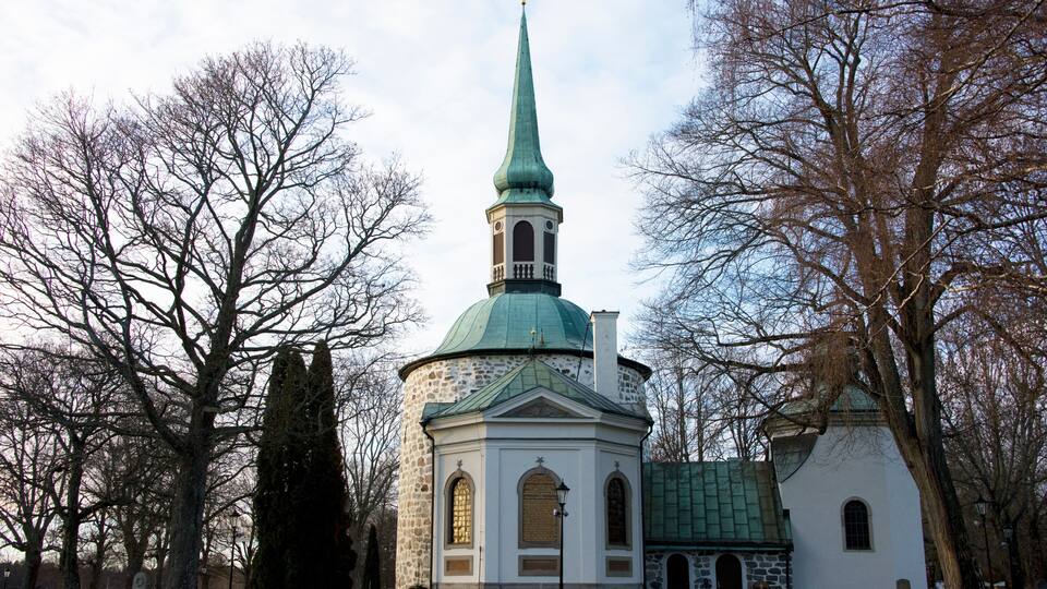A church with a green spire and stone walls, surrounded by leafless trees.