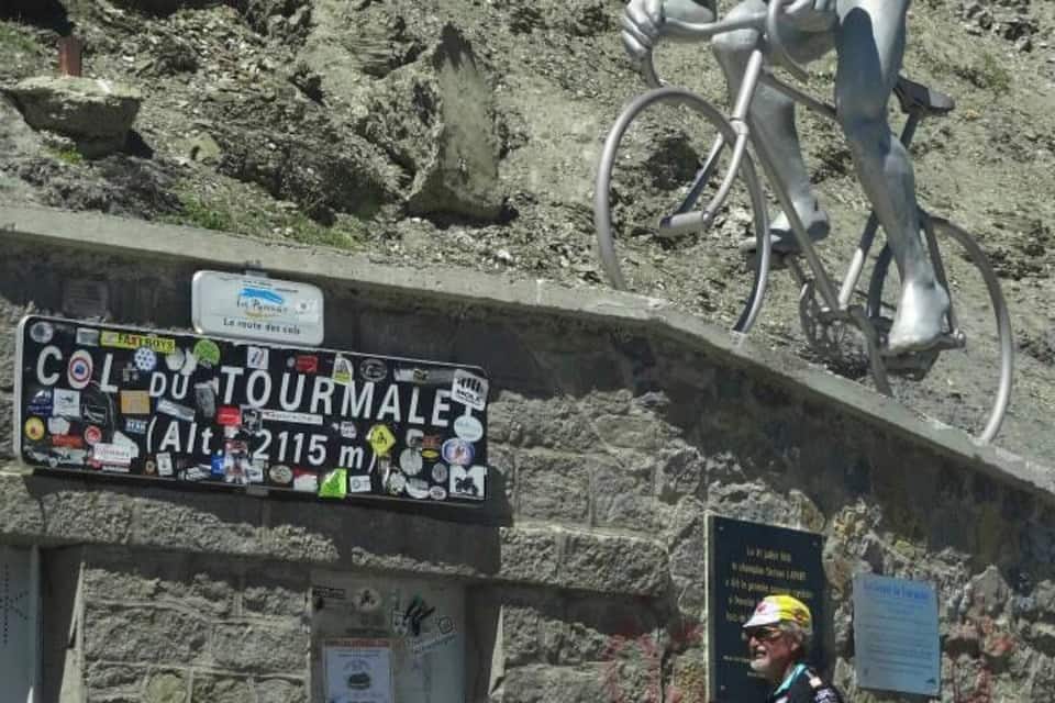 A mountain bike route sign, a cyclist, and a statue of a cyclist on a rocky slope.