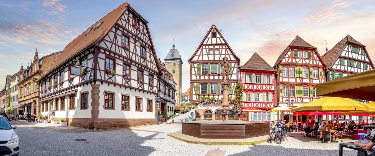 A cobblestone square with traditional half-timbered buildings, a fountain, and outdoor seating areas.