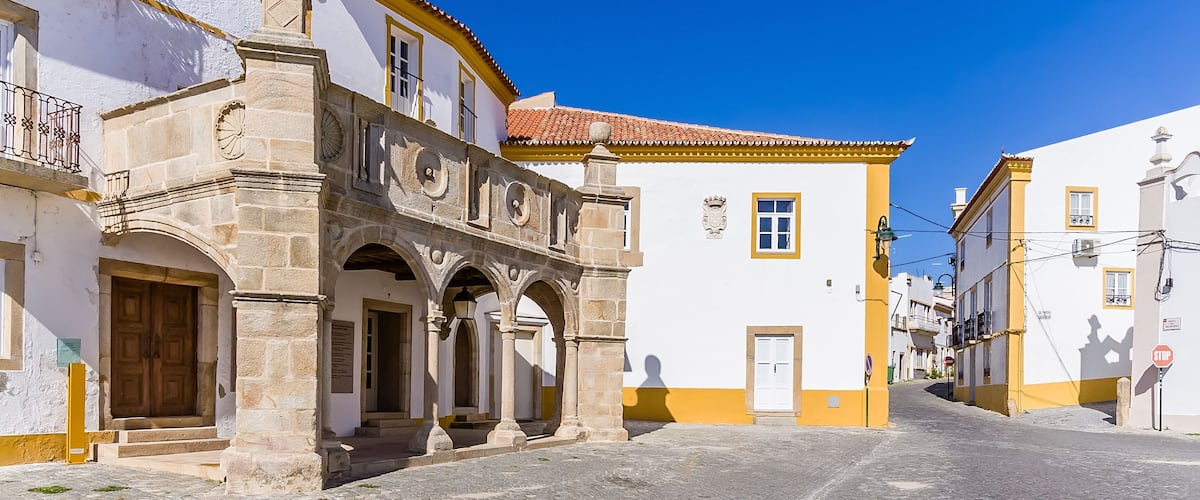 A cobblestone street with historic buildings, a clock tower, and a clear blue sky.
