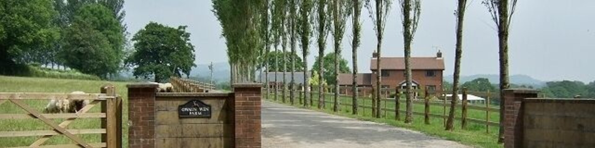Una entrada con portón, un letrero, un sendero de tierra, árboles y un edificio al fondo.