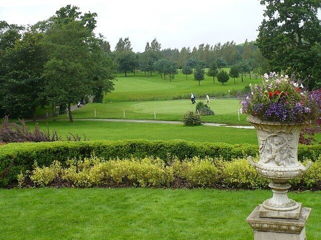 A golf course with a well-manicured lawn, a decorative urn, and a golfer in the distance.