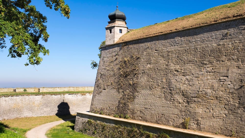 Un mur de pierre surmonté d’une tour de guet, un ciel dégagé et un sentier menant au mur.