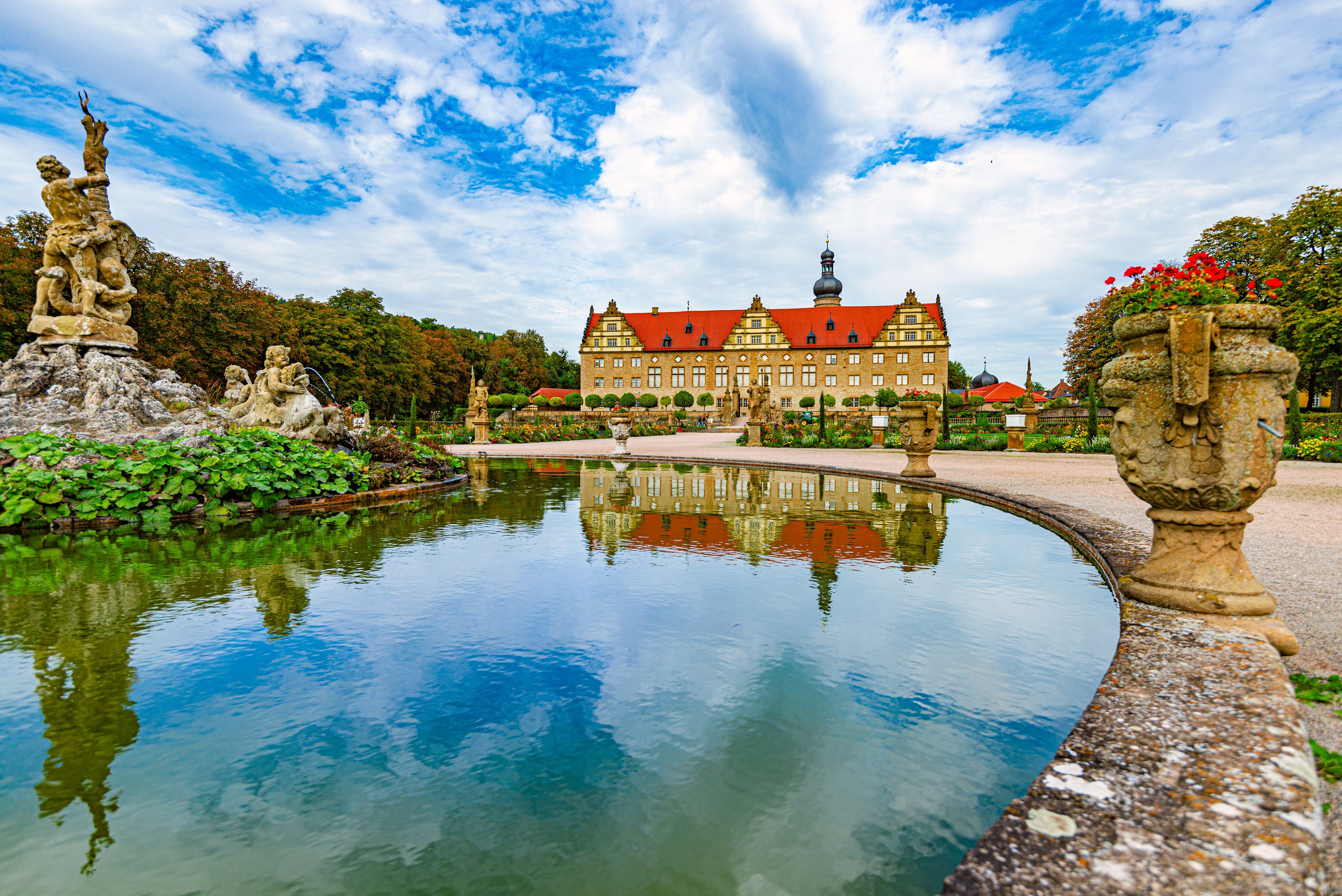 Ein historisches Gebäude mit rotem Dach, ein großer Teich, der die Struktur widerspiegelt, und Statuen rund um den Teich.