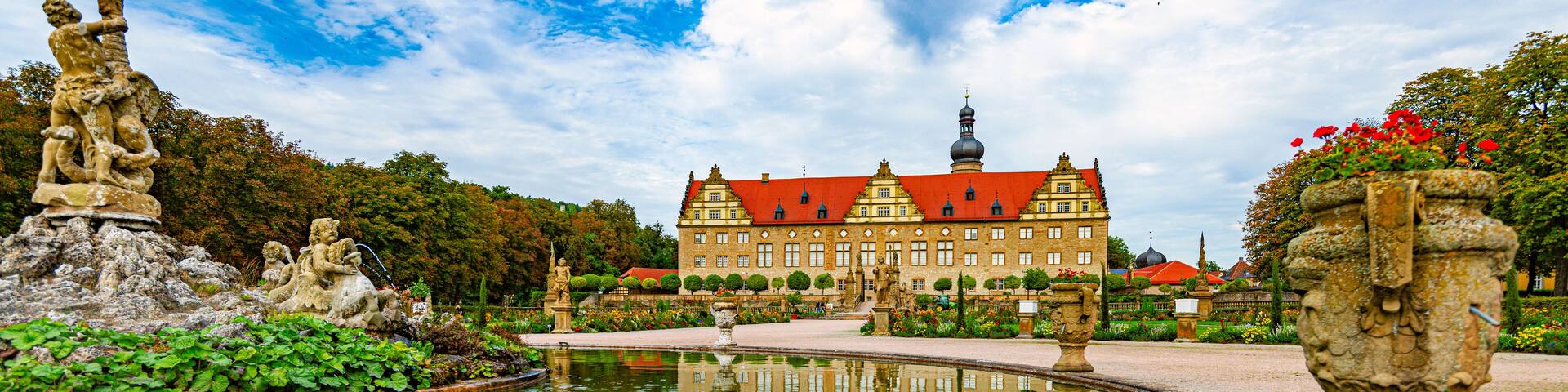 A historic building with a red roof, a large pond reflecting the structure, and statues around the pond.