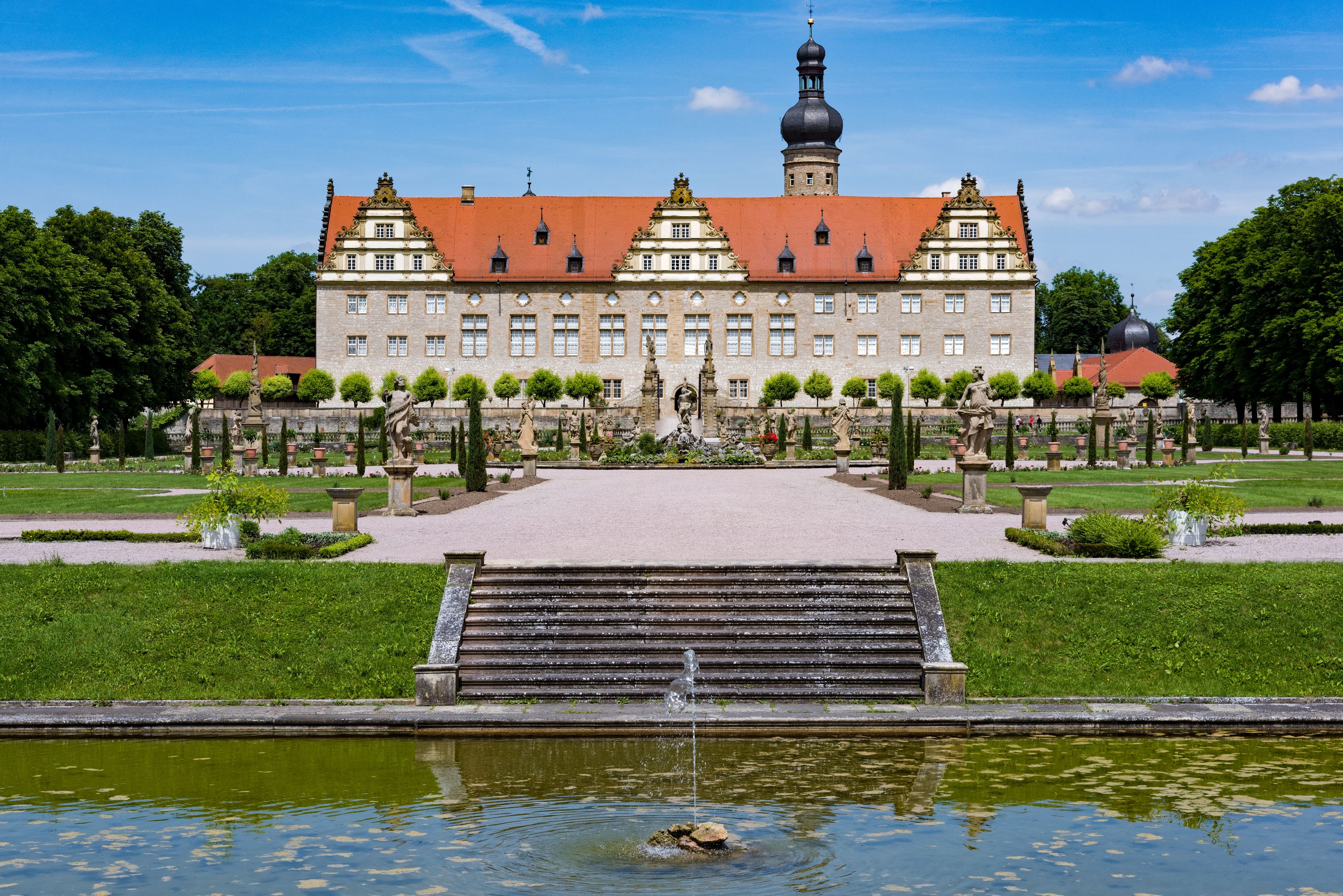 Ein historisches Gebäude mit rotem Ziegeldach, umgeben von einem formalen Garten mit Statuen und einem zentralen Brunnen.