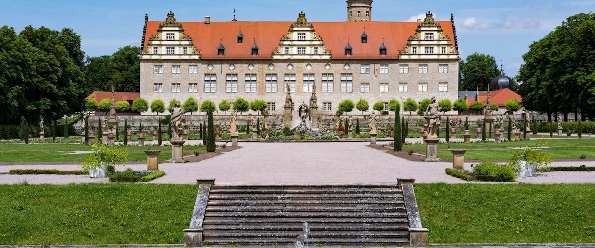 Un bâtiment historique, doté d’un toit recouvert de tuiles rouges, entouré d’un jardin à la française agrémenté de statues et d’une fontaine centrale.