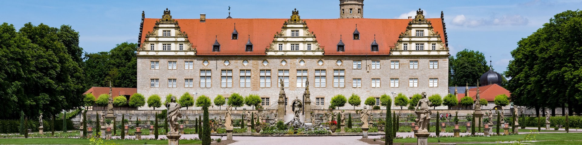 A historic building with a red-tiled roof, surrounded by a formal garden with statues and a central fountain.