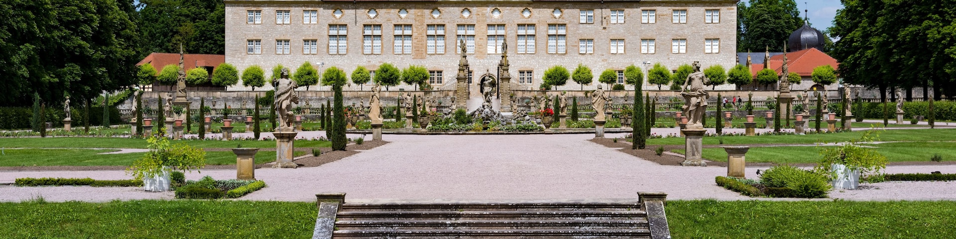 Un edificio histórico con tejado de tejas rojas, rodeado por un jardín formal con estatuas y una fuente central.
