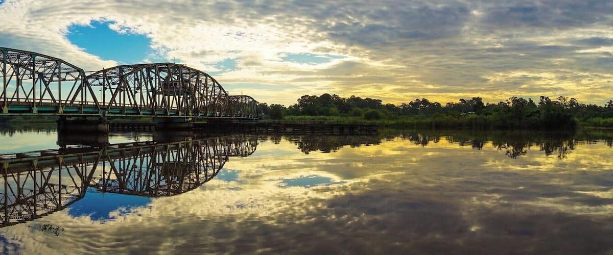 A steel bridge over a calm river with reflections of the sky and trees.