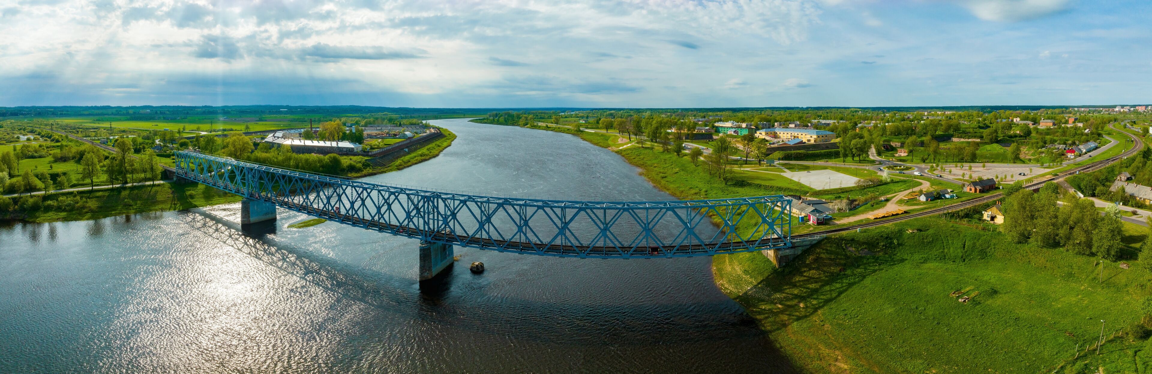 A blue bridge over a river, surrounded by green fields and small buildings.