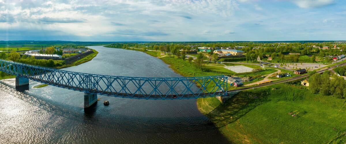 A blue bridge over a river, surrounded by green fields and small buildings.