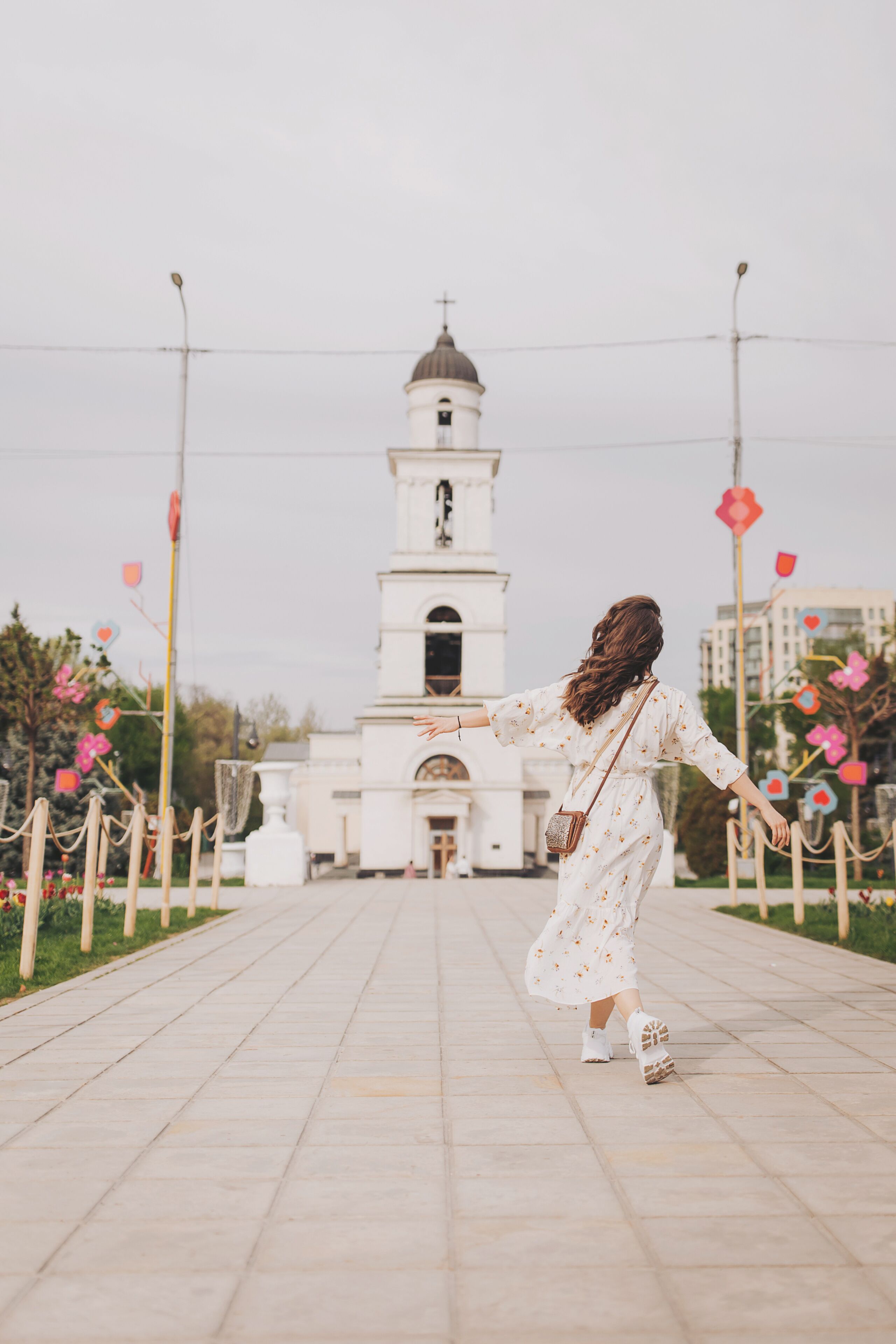 Una persona con un vestido blanco estampado con flores caminando por un sendero pavimentado hacia una iglesia blanca con un campanario.