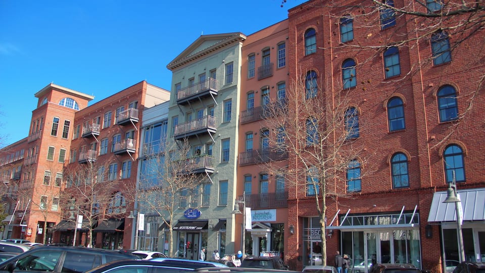 A row of multi-story buildings with commercial storefronts, parked cars, and leafless trees.