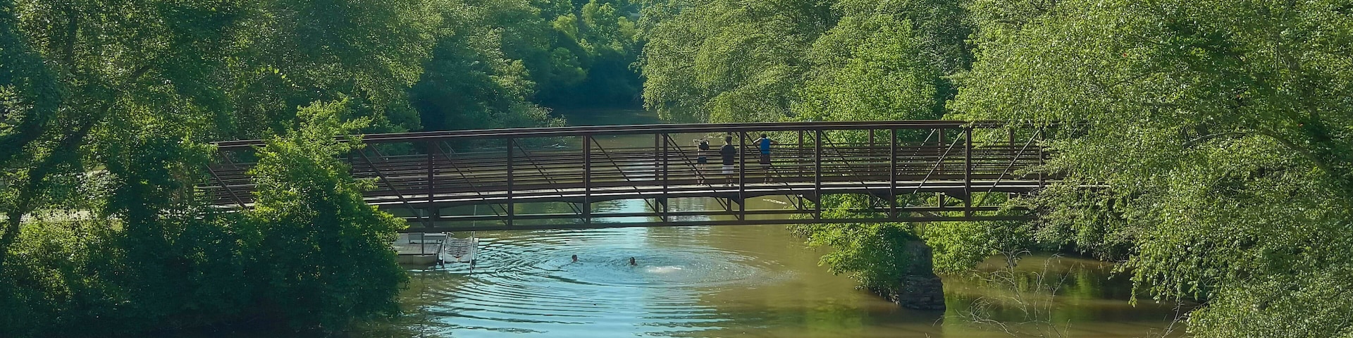 a long metal rust colored bridge over the silk brown waters of Little River with kids diving off the bridge surrounded by lush green trees, grass and plants with blue sky and clouds