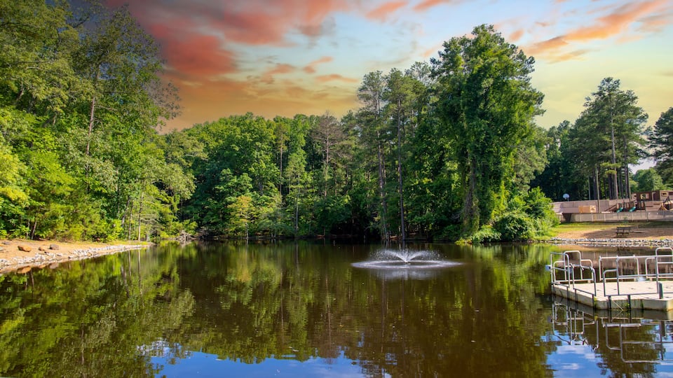 a still lake with a water fountain in the center surrounded by lush green trees and grass reflecting off the water with powerful clouds at sunset at Dupree Park in Woodstock Georgia USA
