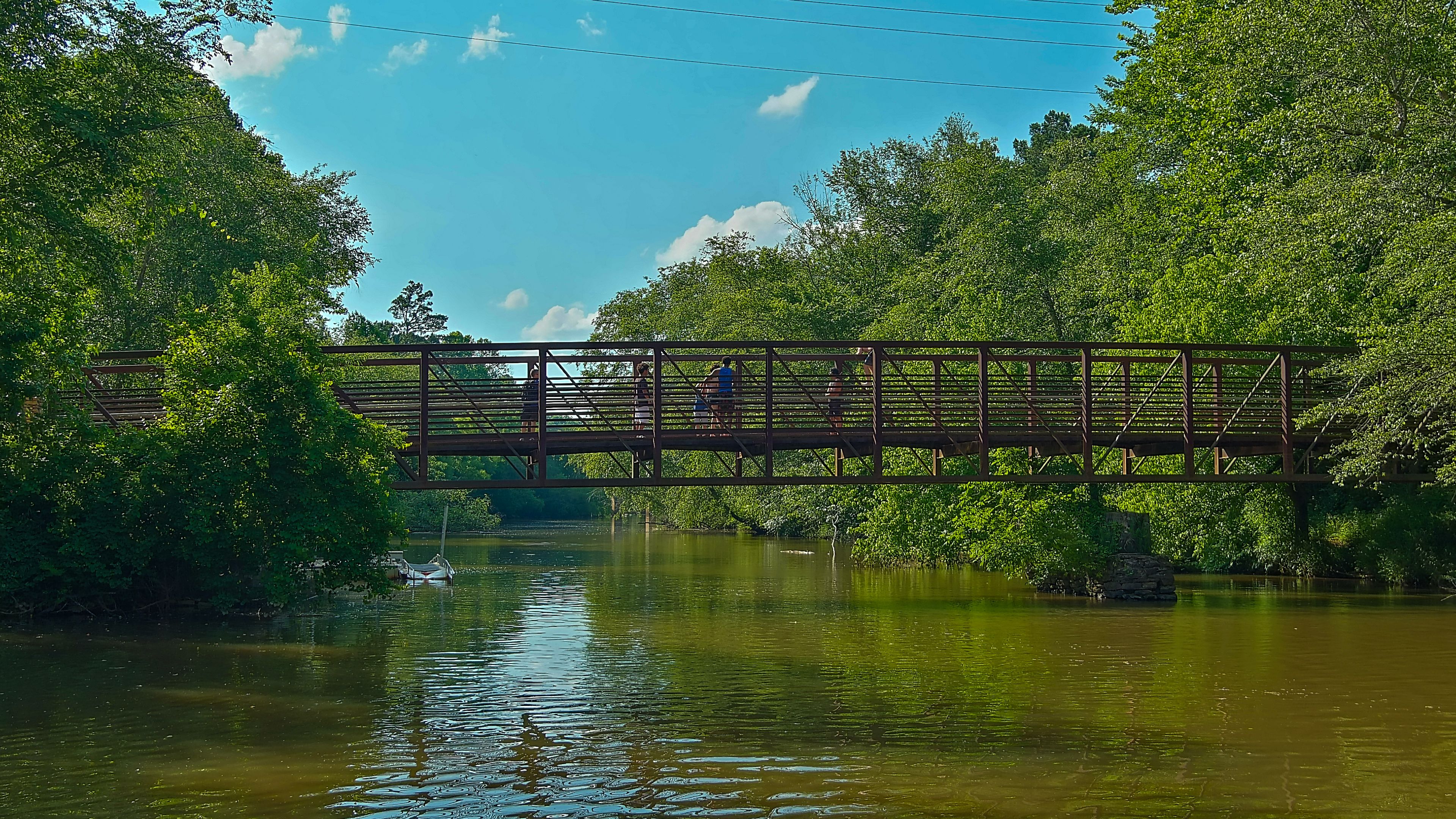 a long metal rust colored bridge over the silk brown waters of Little River with kids diving off the bridge surrounded by lush green trees, grass and plants with blue sky and clouds