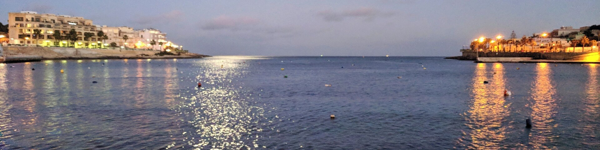 Una ciudad costera al anochecer, con un mar tranquilo que refleja las luces de la ciudad.