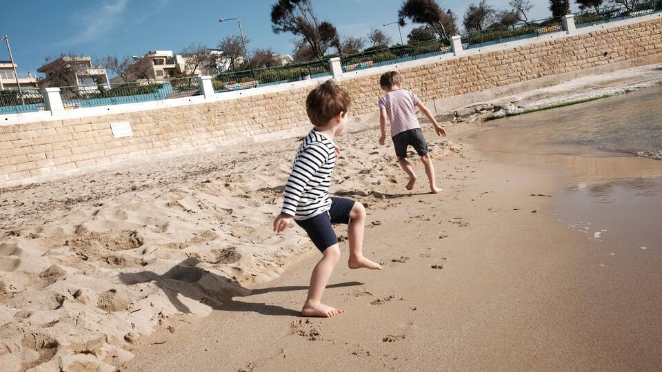 Two children playing on a sandy beach with a stone wall and a fence in the background.