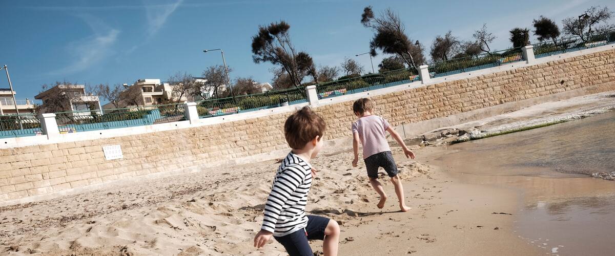 Two children playing on a sandy beach with a stone wall and a fence in the background.