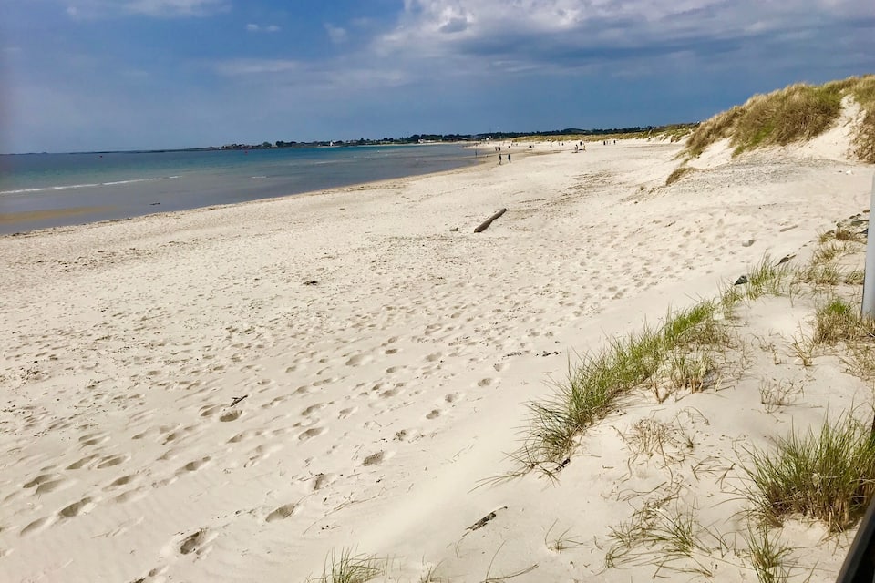 Platja sorrenca amb petjades i herba escassa, mar tranquil·la i cel serè.
